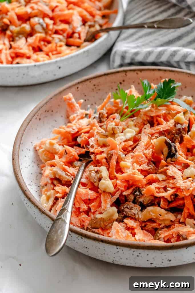 A vibrant bowl of homemade Carrot Raisin Salad, showcasing bright orange shredded carrots, a mix of dark and golden raisins, and chopped walnuts, all tossed in a creamy yogurt dressing. The salad is presented in a white ceramic bowl, ready to be served.