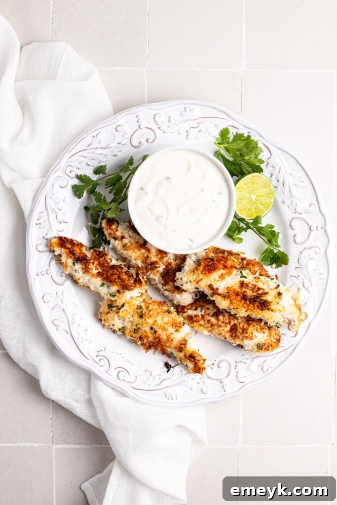Golden Crispy Baked Coconut Chicken Tenders Kids Love 5 Crispy Coconut Chicken Tenders on a white plate with a bowl of cilantro lime dipping sauce on the side.