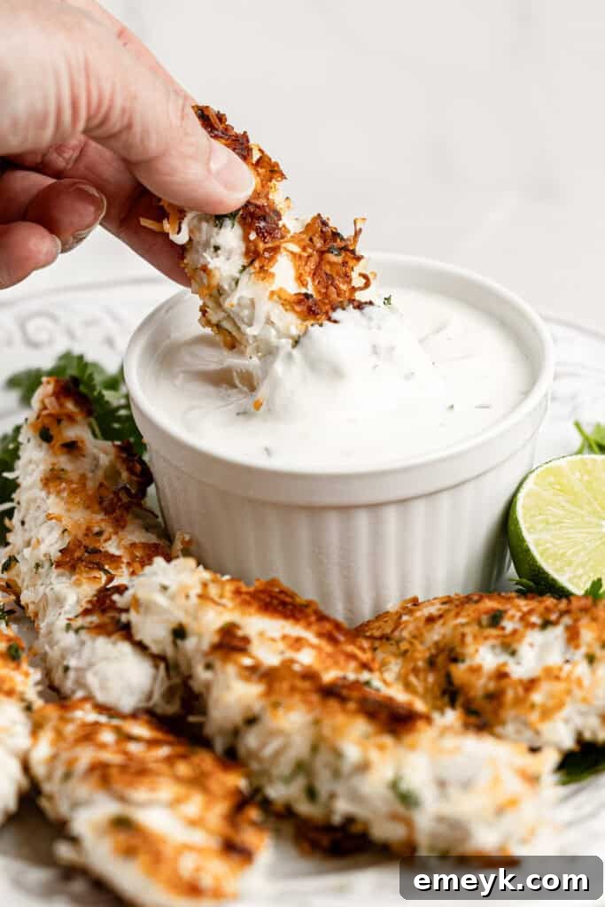 Golden Crispy Baked Coconut Chicken Tenders Kids Love 9 A crispy coconut chicken tender being dipped into a bowl of creamy aioli.