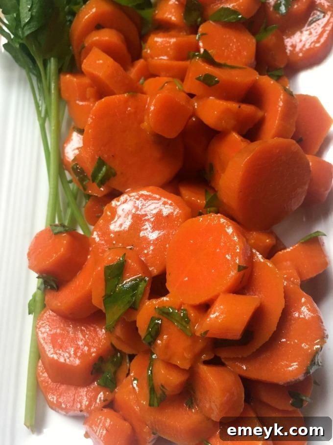 A skillet filled with vibrant orange carrots cooking in broth and butter, with a spoonful of honey being added.