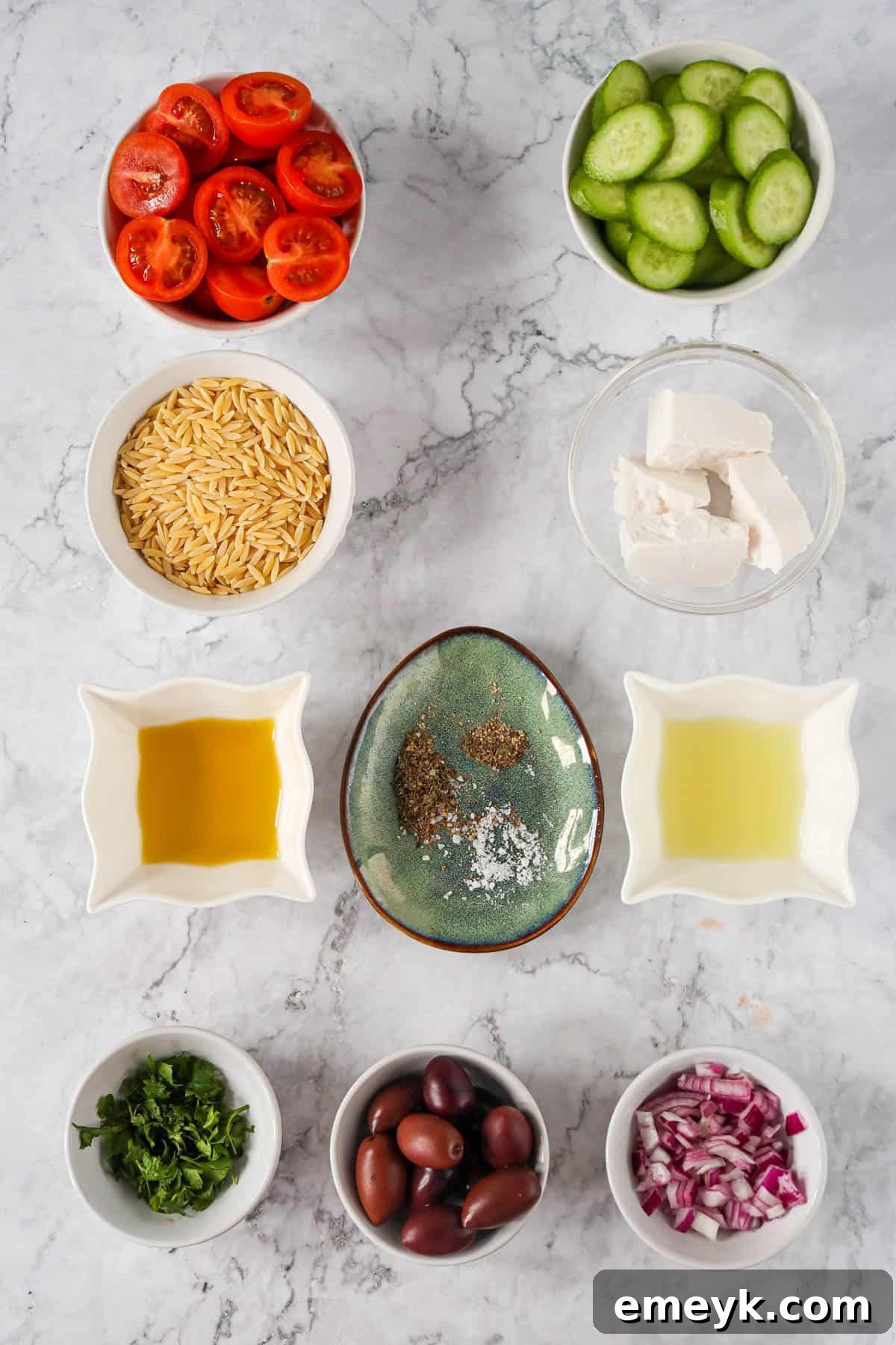 A flat lay of all the fresh ingredients for Greek Orzo Pasta Salad, including cherry tomatoes, cucumbers, feta, and a bowl of orzo.