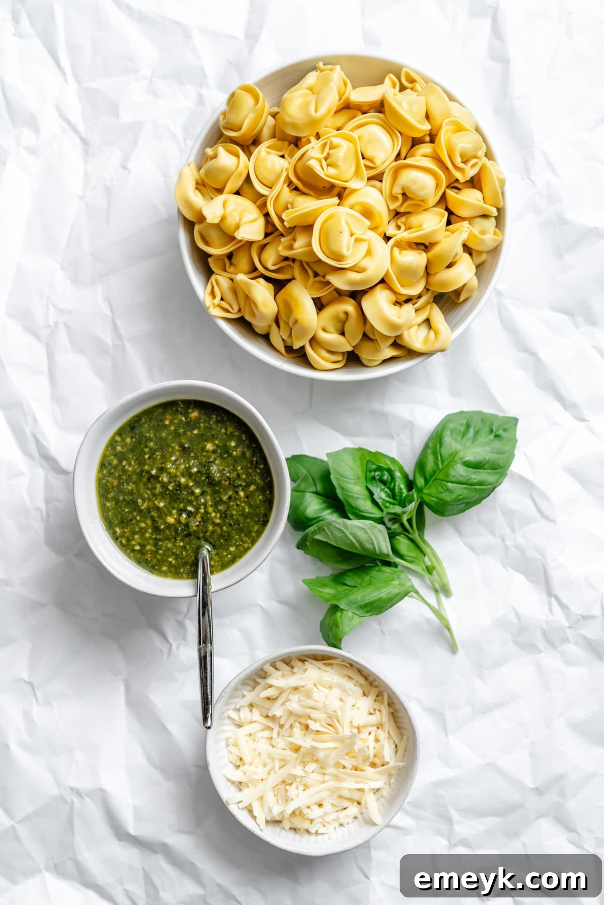 Small bowls containing the three main ingredients for pesto tortellini: fresh tortellini, grated Parmesan cheese, and a vibrant green pesto sauce.