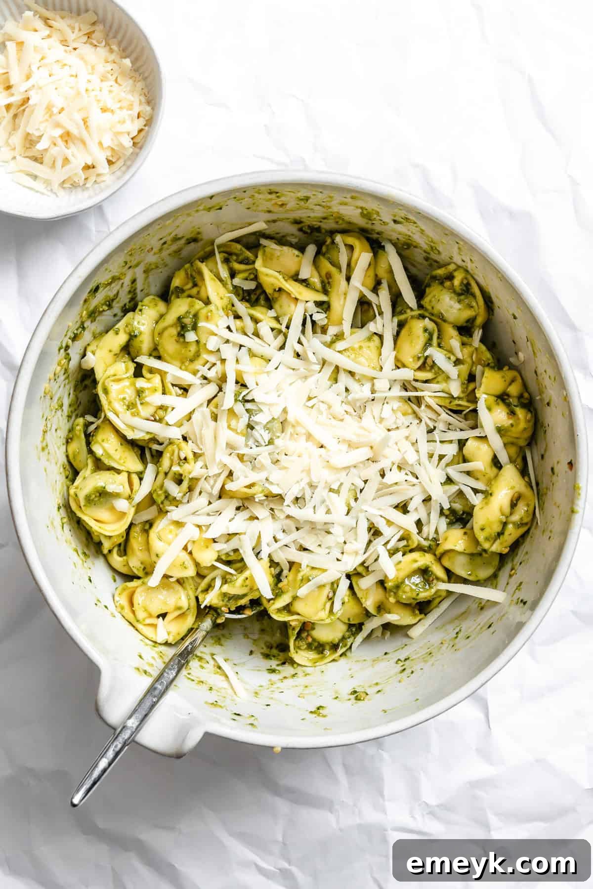 Pesto-coated tortellini generously mixed with Parmesan cheese in a large glass mixing bowl.
