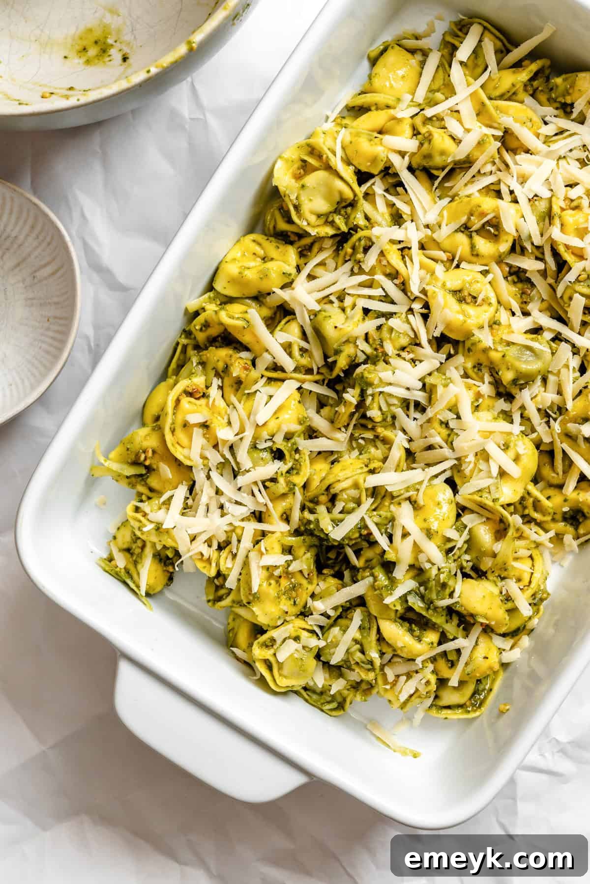 Pesto tortellini spread in a baking dish, topped with a generous layer of Parmesan cheese, ready for the oven.