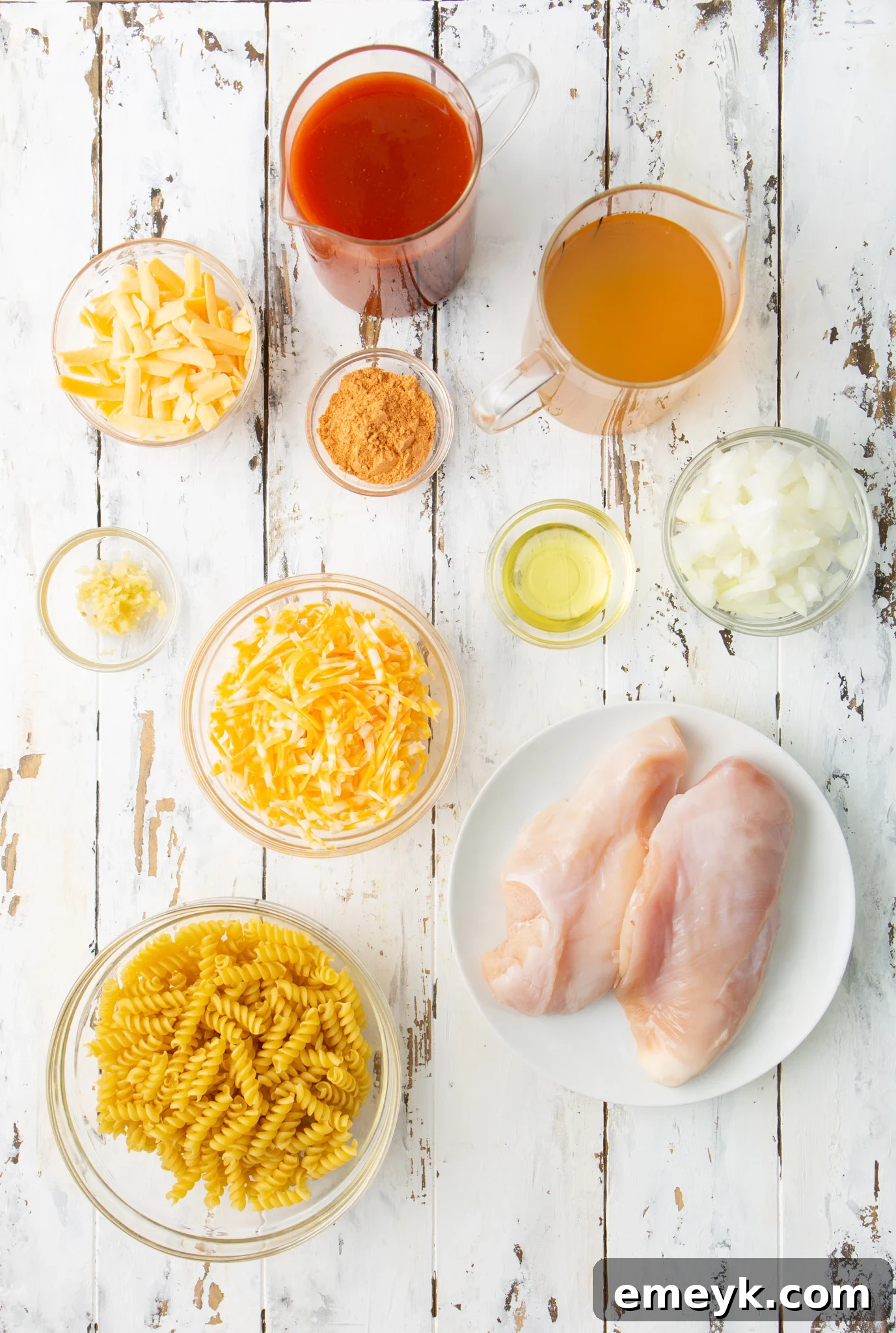 A flat lay of fresh ingredients including rotini pasta, chicken breasts, sweet onion, garlic cloves, and blocks of cheese, ready for One Pot Chicken Enchilada Pasta.