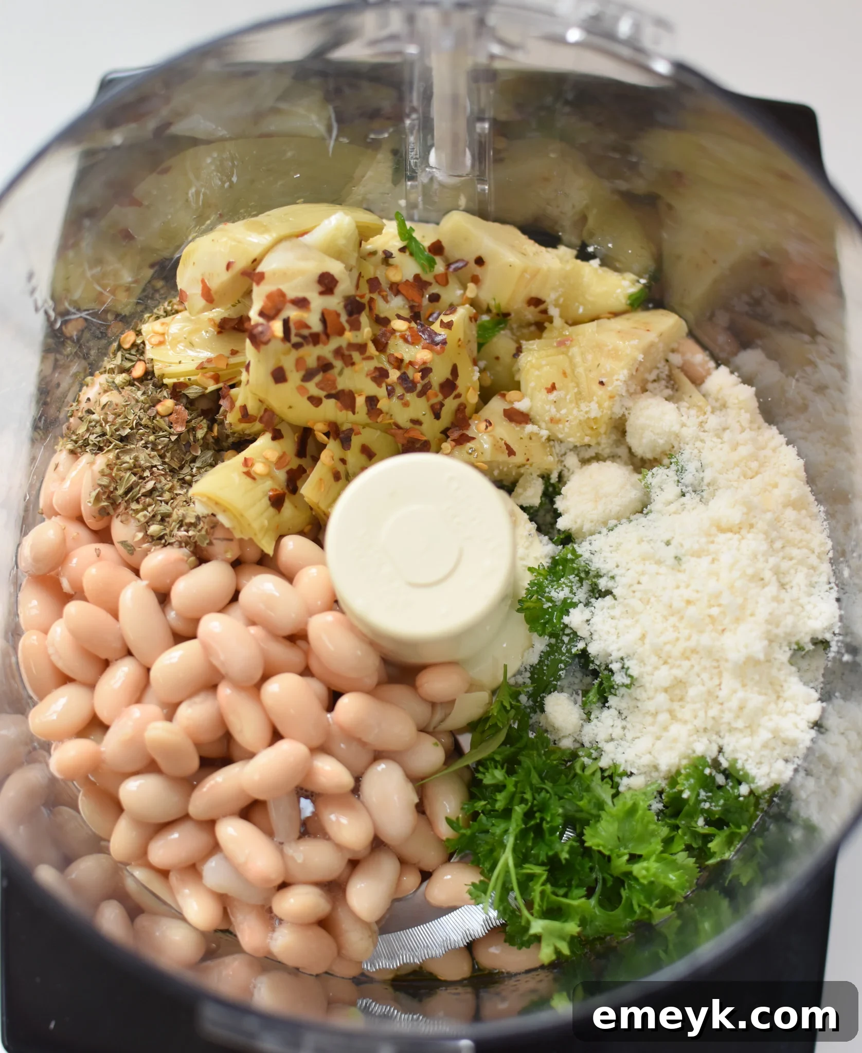 All the core ingredients for White Bean Artichoke Dip, including cannellini beans, artichoke hearts, garlic, and Parmesan cheese, placed inside a food processor ready for blending.