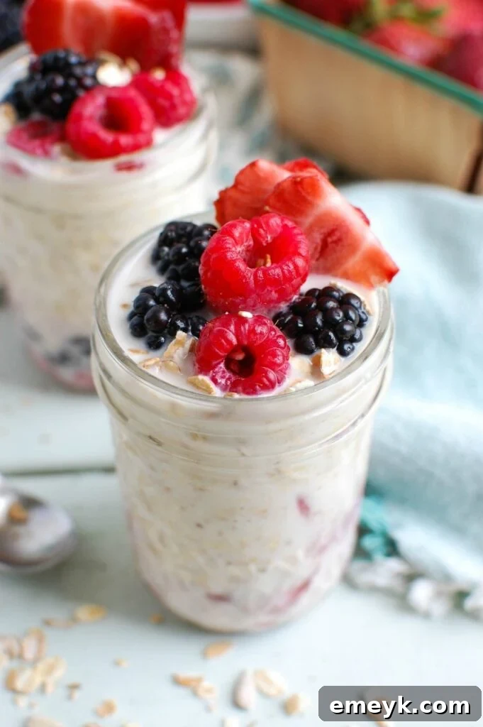 A close-up of a mason jar filled with colorful Overnight Triple Berry Oatmeal, showcasing layers of oats, milk, Greek yogurt, and fresh blueberries, raspberries, and sliced strawberries. It's ready to be stored in the fridge for an easy breakfast.