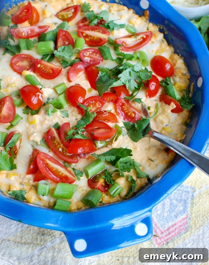 Side view of Baked Sriracha Sweet Corn Dip showing layers of melted cheese, corn, and fresh garnishes, with a serving spoon and chips nearby.