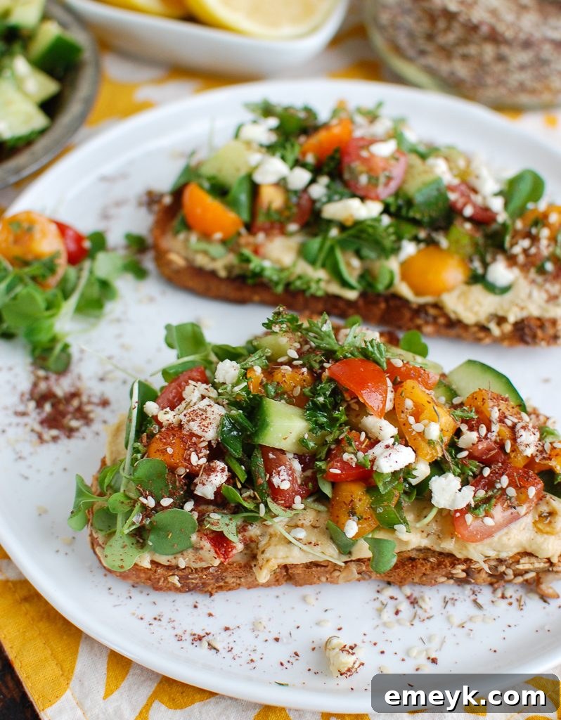 Two pieces of Mediterranean Hummus Toast with Za'atar on a plate, topped with fresh tomatoes, cucumbers, herbs, and feta cheese.