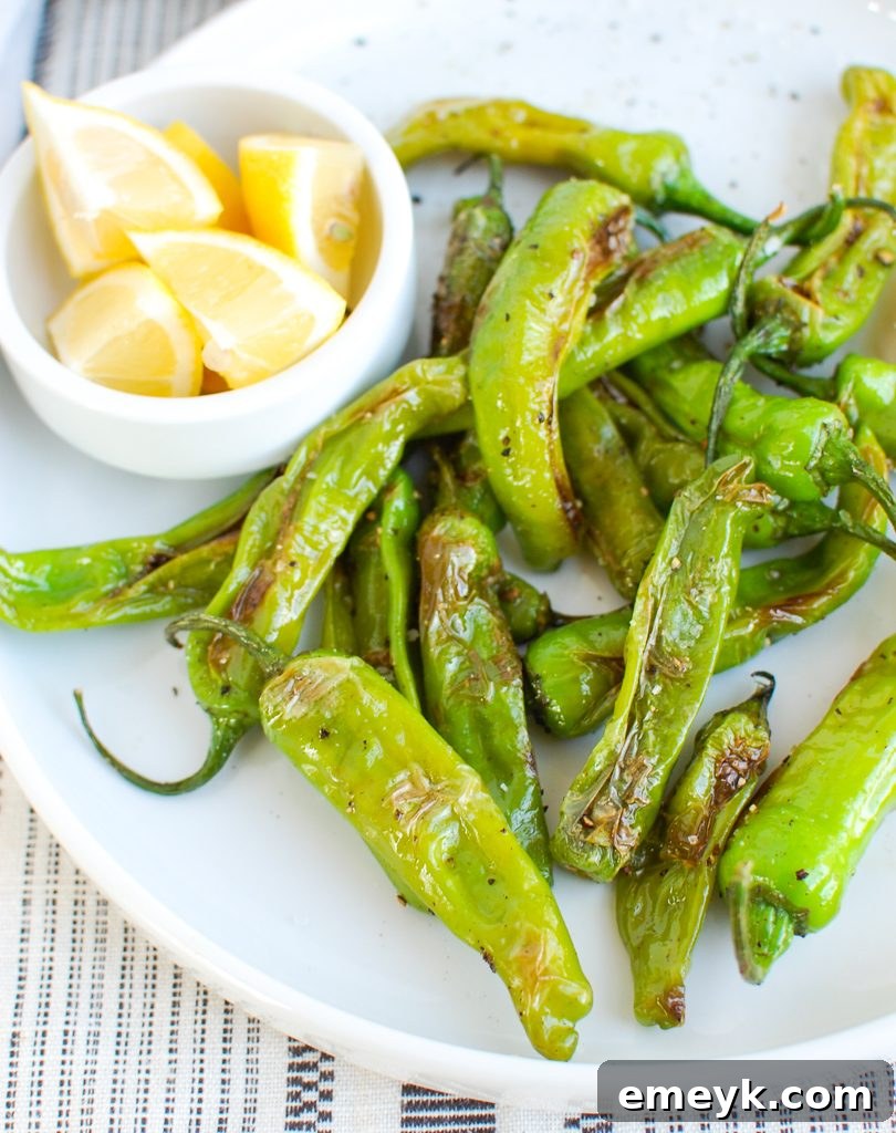 A serving of cooked Shishito Peppers garnished with lemon wedges, on a rustic board
