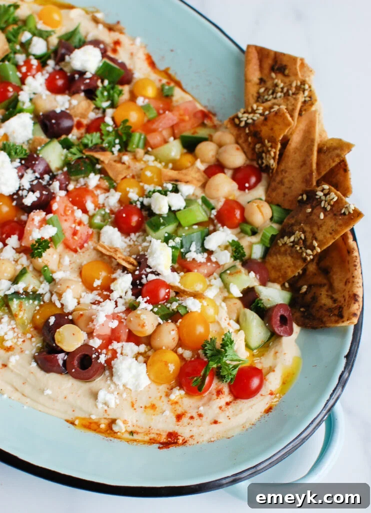 Mediterranean Hummus Board 2 Hummus Platter on a teal plate, beautifully garnished with fresh Mediterranean toppings including olives, feta, tomatoes, and herbs.