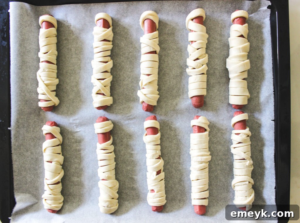 Pharaoh Franks 5 Several hot dogs individually wrapped with strips of puff pastry, resembling mummies, laid out on a cutting board before baking.