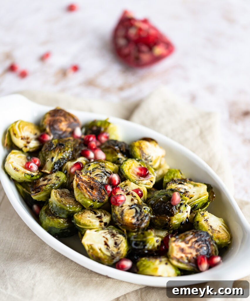 Close-up of Roasted Balsamic Brussels Sprouts in a white serving bowl, richly coated in glaze and adorned with bright red pomegranate arils.