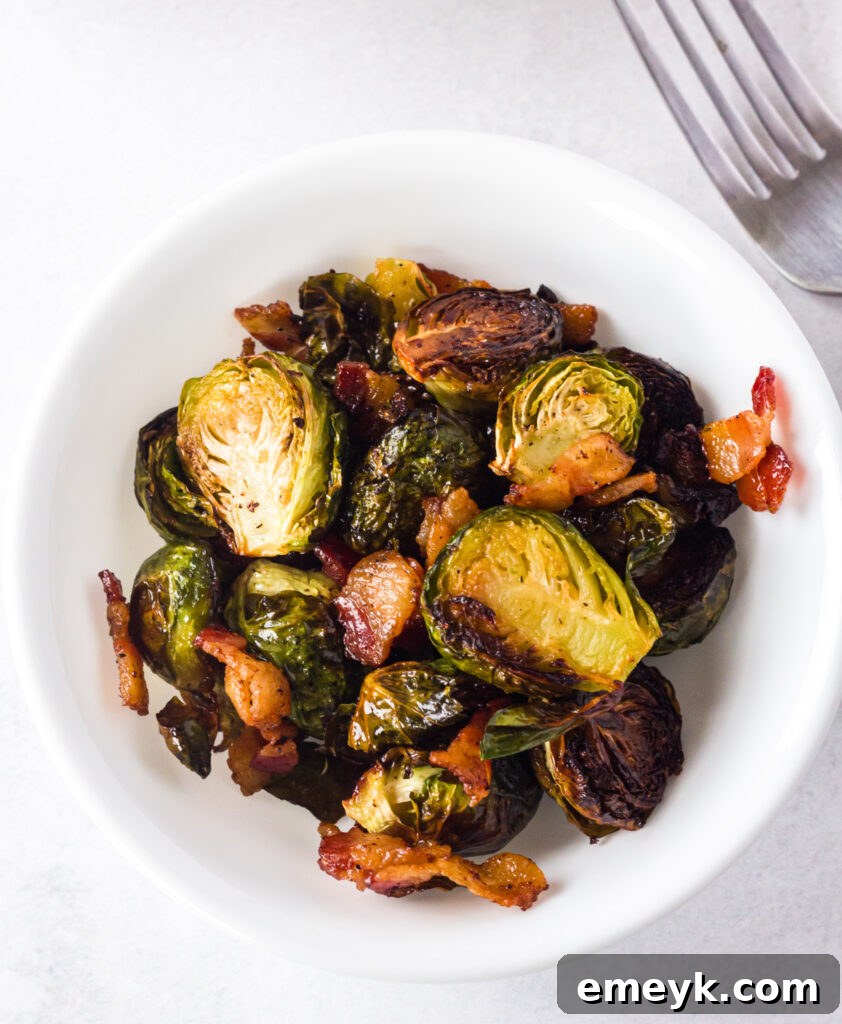 A fork piercing a piece of roasted Brussels sprout and bacon in a white bowl, showcasing the crispy texture.