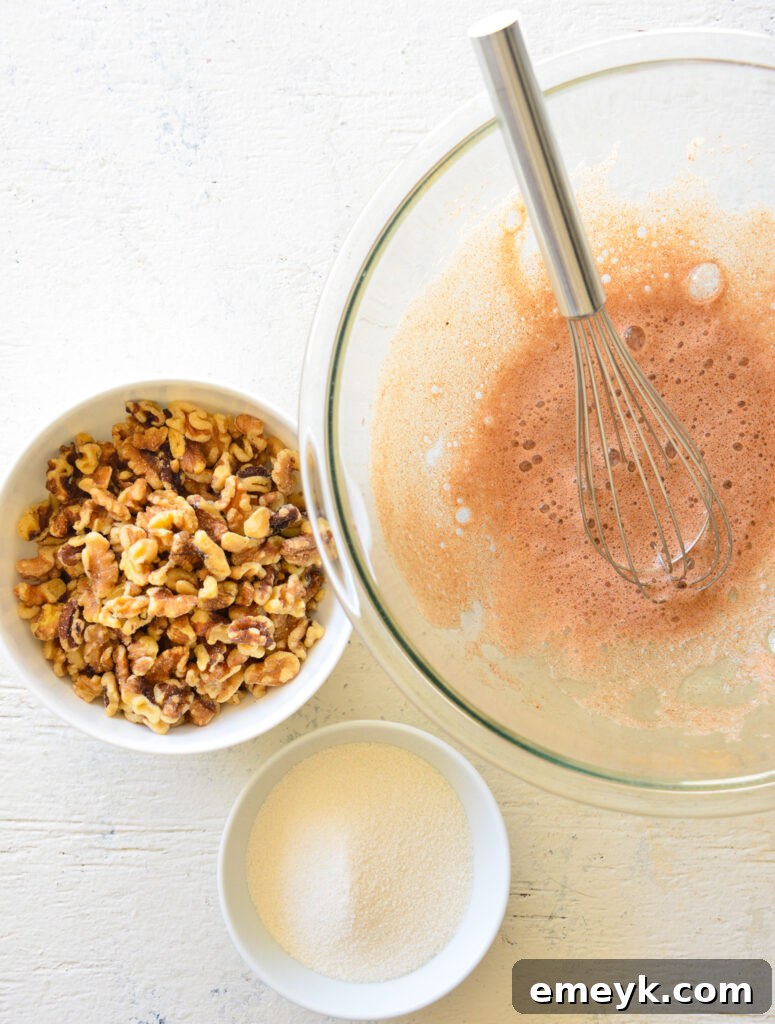 A hand whisking together the egg white, sugar, vanilla, cinnamon, salt, and water in a glass bowl, creating a foamy mixture for the candied walnuts.