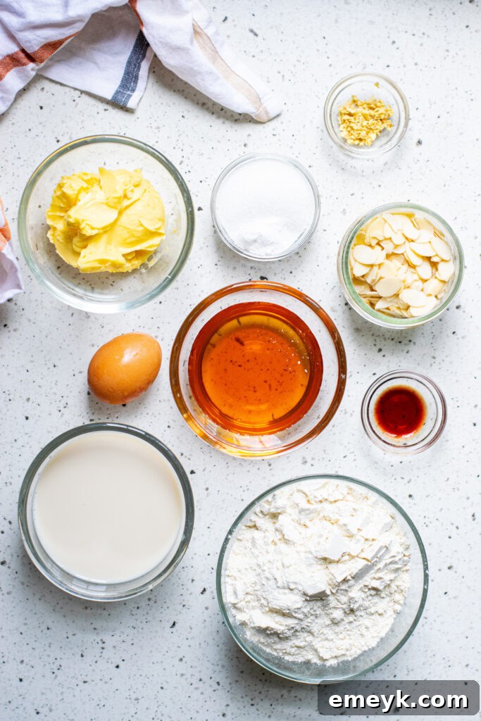 Essential ingredients for a delicious Honey Almond Cake laid out on a rustic wooden surface, including flour, baking powder, fresh ginger, sugar, sea salt, butter, egg, honey, whole milk, vanilla extract, and slivered almonds, ready for baking.