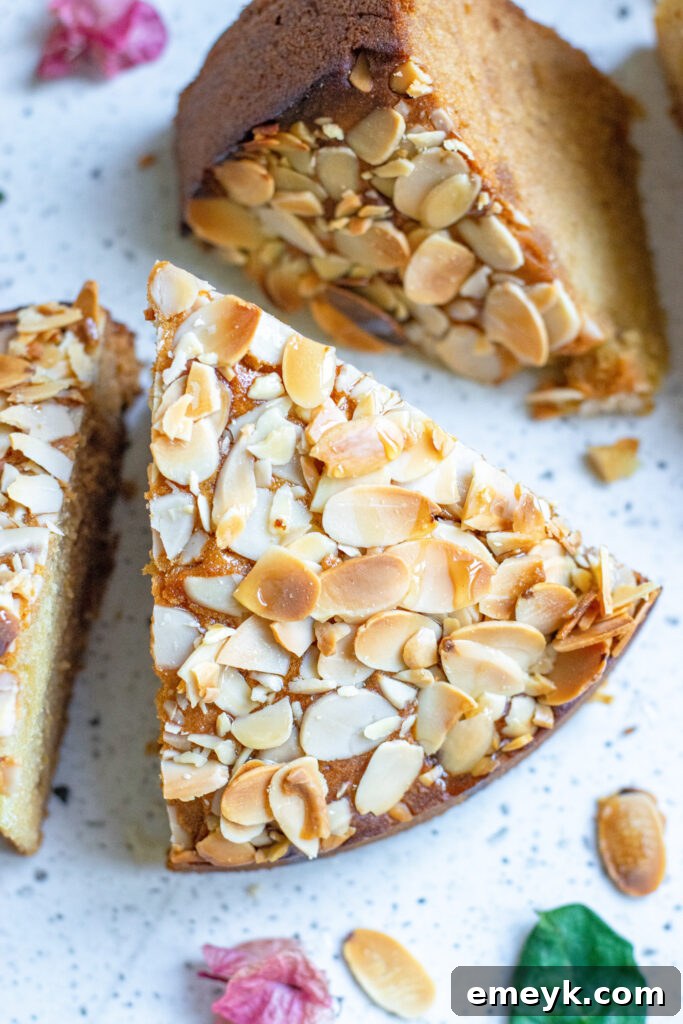A delectable slice of Honey Almond Cake, showcasing its golden crust, generous topping of toasted slivered almonds, and moist interior, served on a white plate with a fork.
