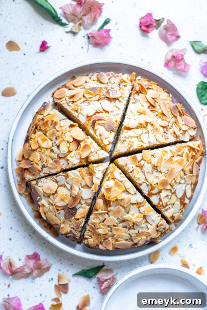 An inviting overhead view of a whole Honey Almond Cake, beautifully topped with golden-brown slivered almonds, presented on a wooden board, with a few slices already cut, revealing its moist interior.