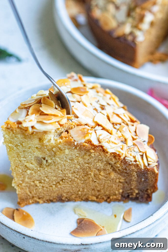 A close-up of a slice of Honey Almond Cake on a plate, with a fork poised to take a bite, highlighting the moist interior and crunchy almond topping.