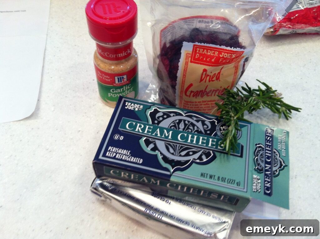 Key ingredients for making Rosemary Cranberry Spread laid out on a cutting board: cream cheese, dried cranberries, fresh rosemary sprigs, and garlic powder.