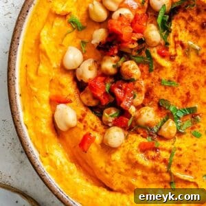 Close-up of creamy Roasted Red Pepper Hummus with a rich red hue, garnished with a sprinkle of paprika and a fresh mint leaf.
