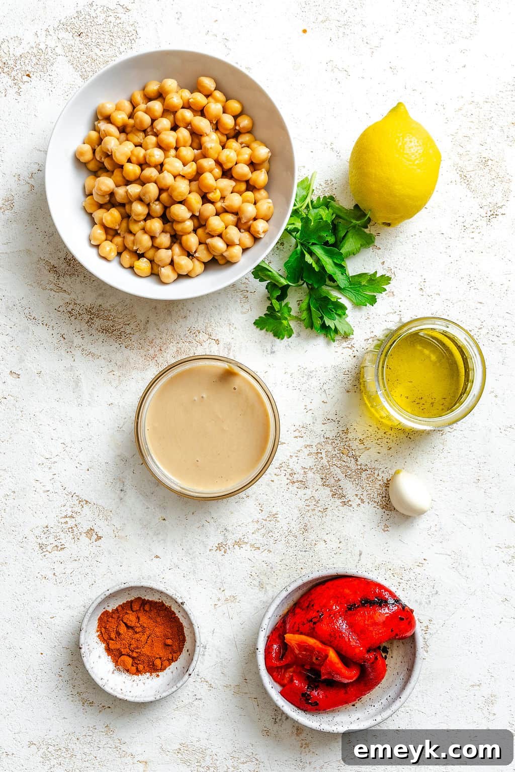 Vibrant Red Pepper Hummus 3 A carefully arranged collection of fresh ingredients for homemade Roasted Red Pepper Hummus, including canned chickpeas, bright lemons, a jar of tahini, garlic cloves, extra virgin olive oil, and vibrant jarred roasted red peppers, all laid out on a rustic surface.
