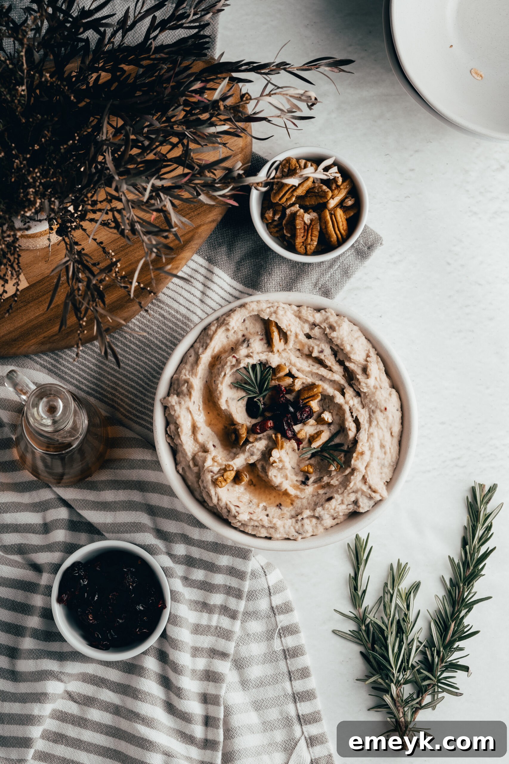 An overhead shot of White Bean Hummus with Cranberry, garnished beautifully with a sprig of fresh rosemary, dried cranberries, and chopped pecans, presented in a rustic white bowl.