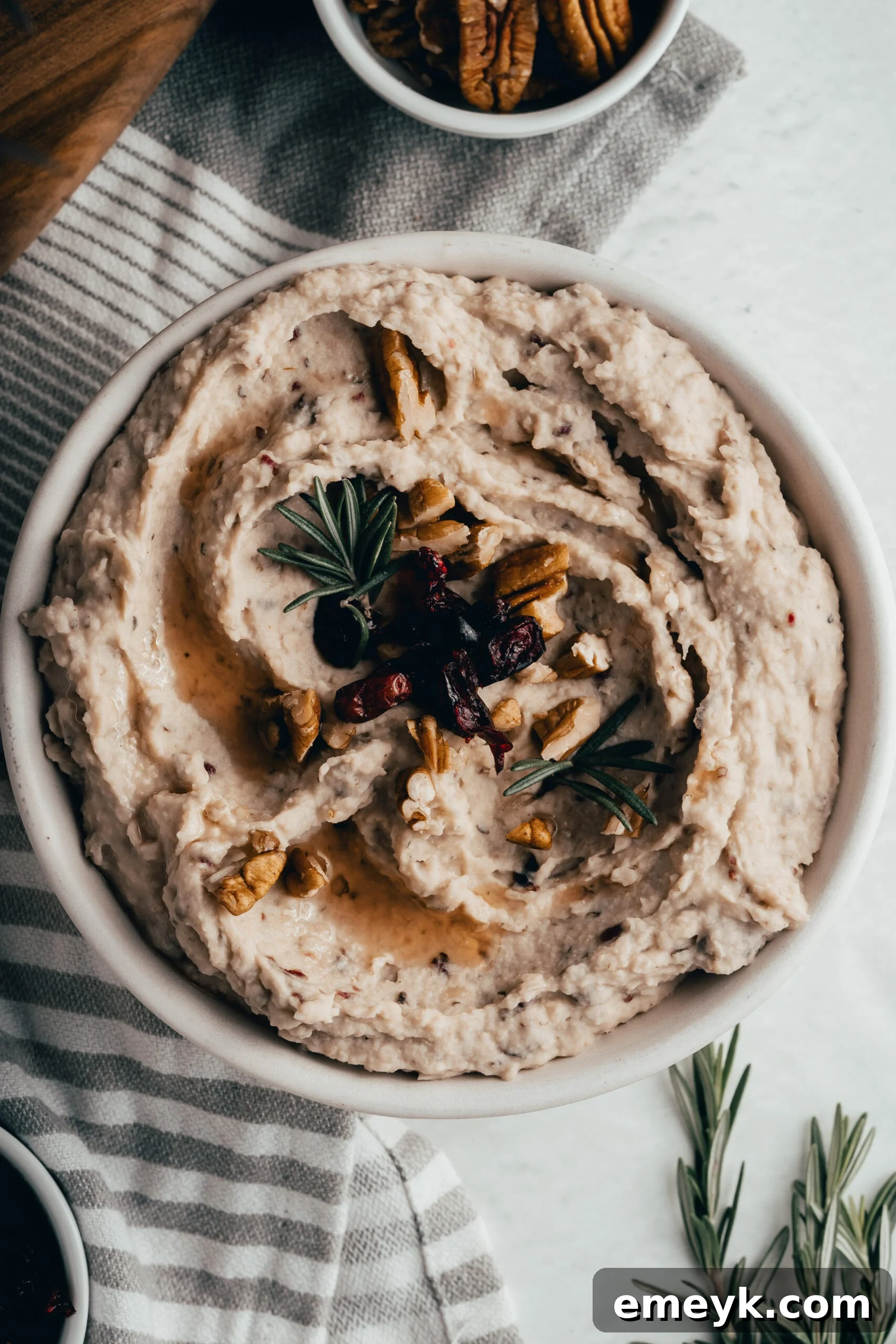 A festive serving of White Bean Hummus with Cranberry, beautifully garnished with a sprig of fresh rosemary, dried cranberries, and chopped pecans.