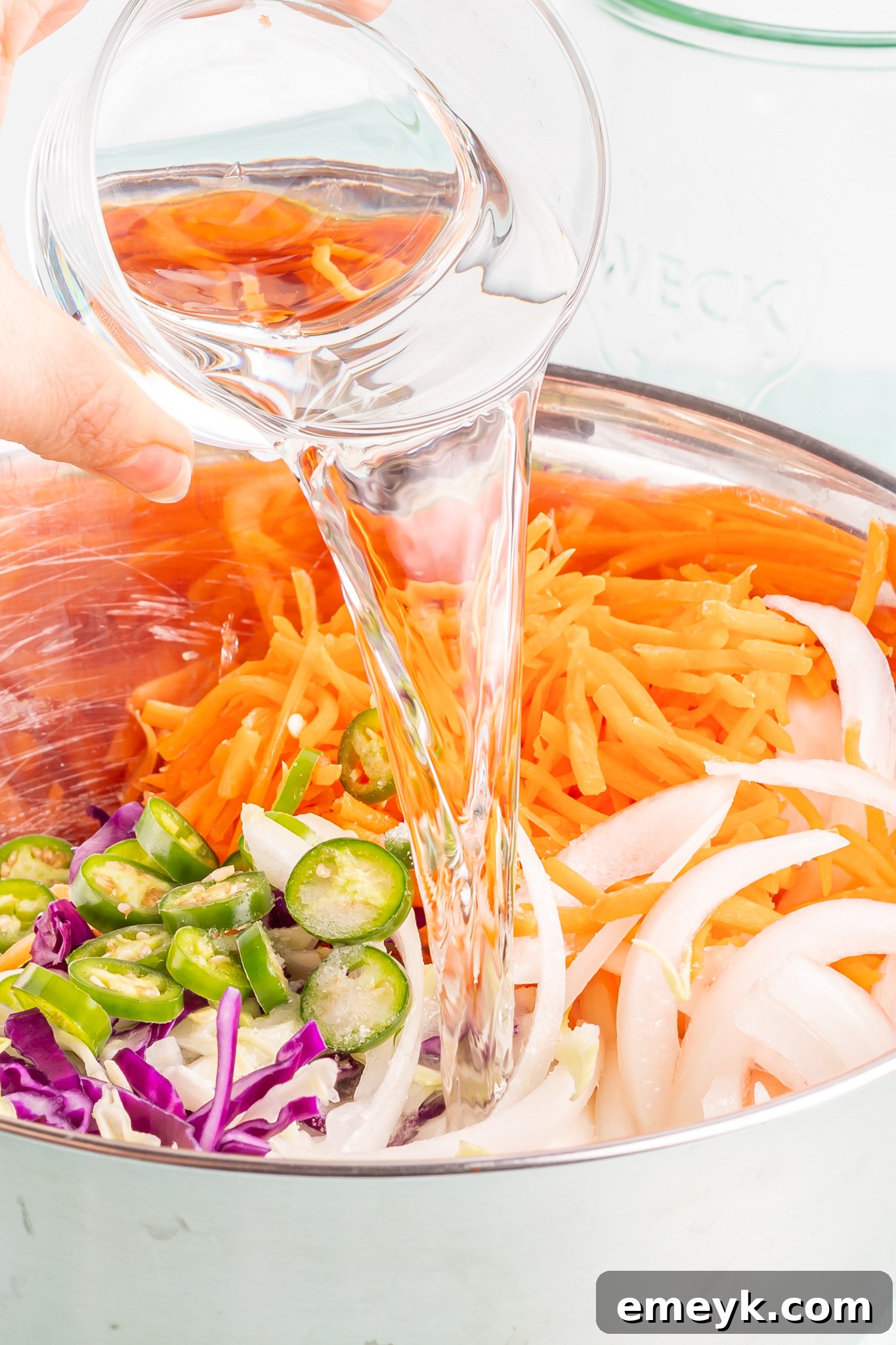 An overhead shot showing white vinegar being poured over the mixed vegetables in a bowl during the Curtido preparation process.