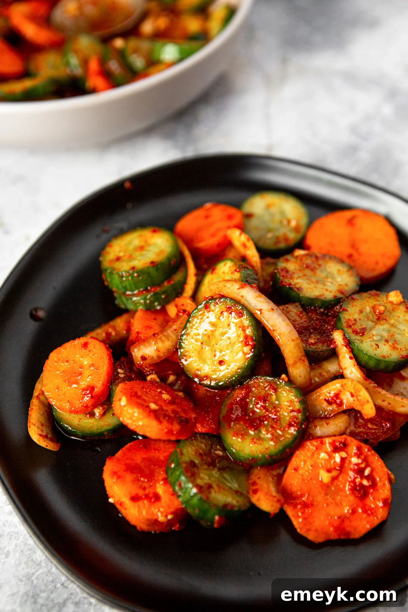A dark bowl filled with vibrant Quick Korean Cucumber Kimchi, showcasing the crisp cucumbers, carrots, and onions against a dark background, garnished with sesame seeds.
