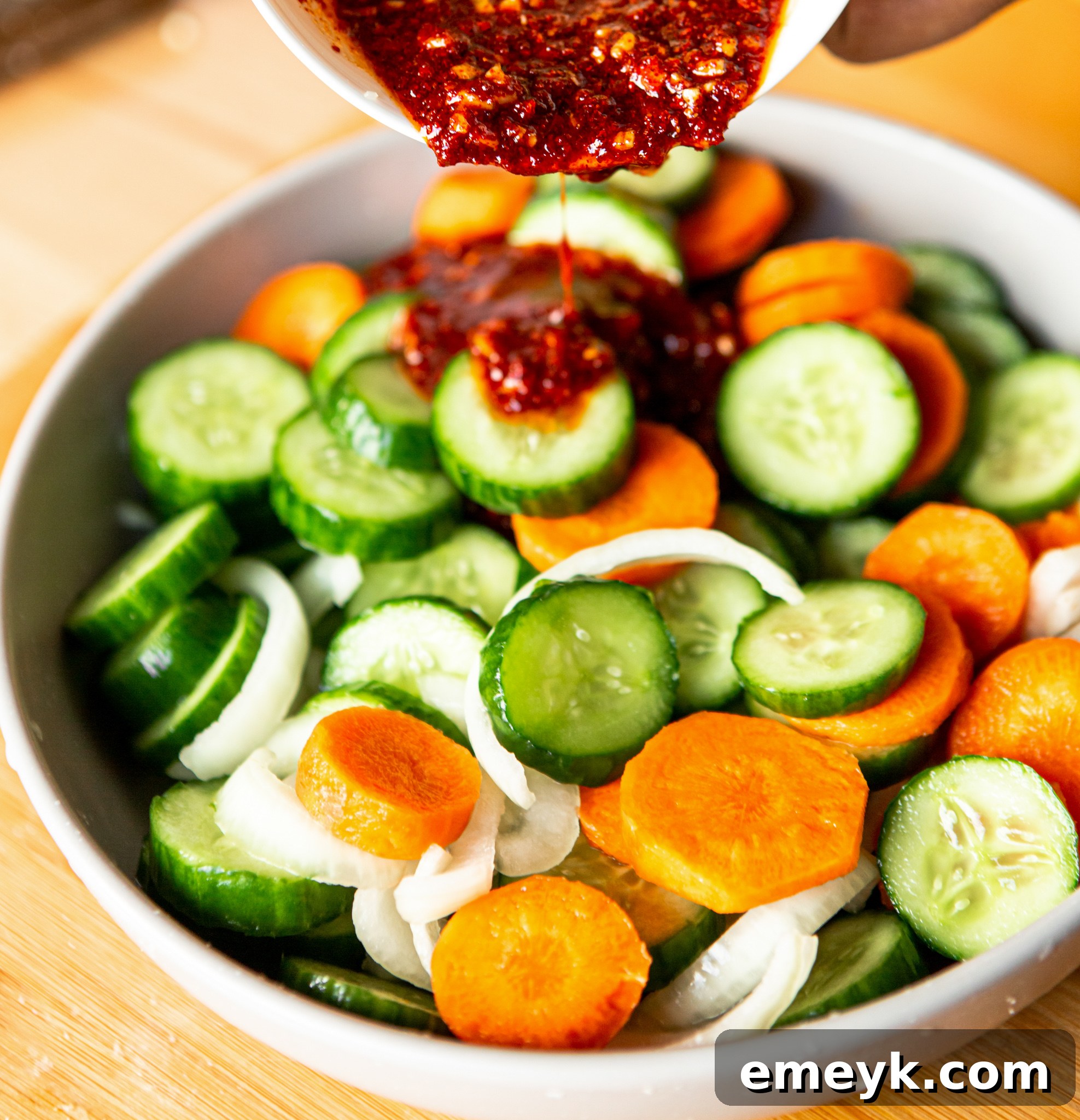 A close-up shot of the kimchi sauce being poured over the freshly rinsed and dried cucumber, carrot, and onion slices in a bowl, just before mixing.