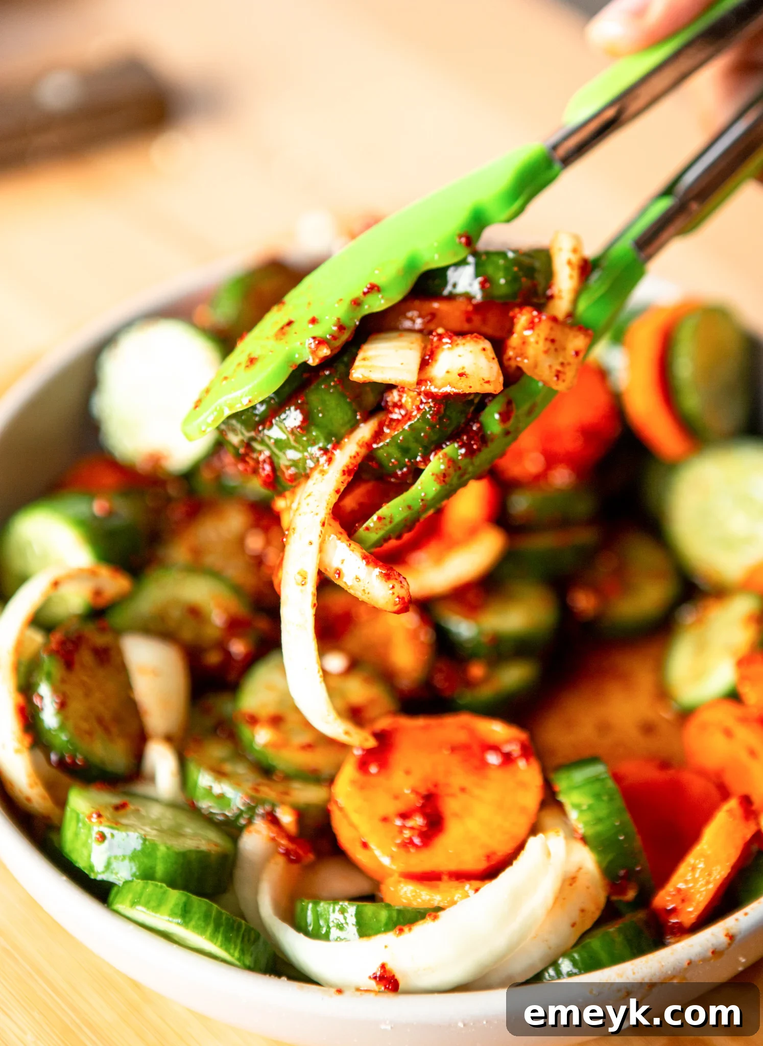 A pair of tongs gently tossing the Quick Korean Cucumber Kimchi in a bowl, ensuring all vegetables are evenly coated with the spicy sauce.