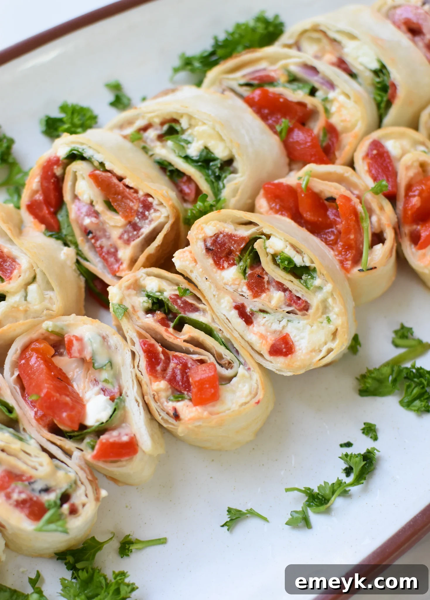 Close-up of Mediterranean Pinwheels artfully arranged on a white plate, showcasing their colorful layers of hummus, feta, roasted red peppers, and arugula. 