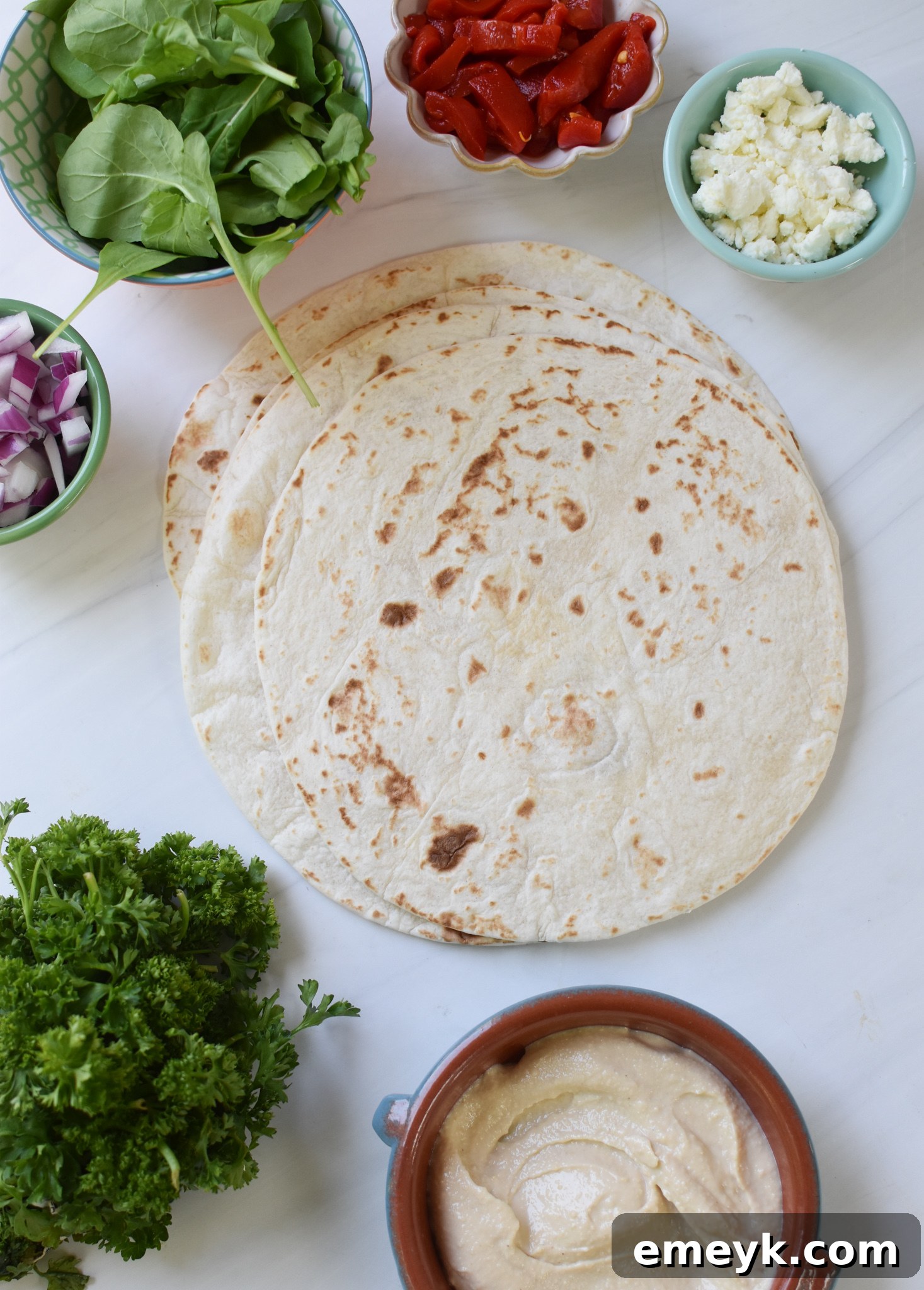 Individual ingredients for Mediterranean Pinwheels laid out: tortillas, hummus, roasted red peppers, feta cheese, arugula, red onion, and parsley.