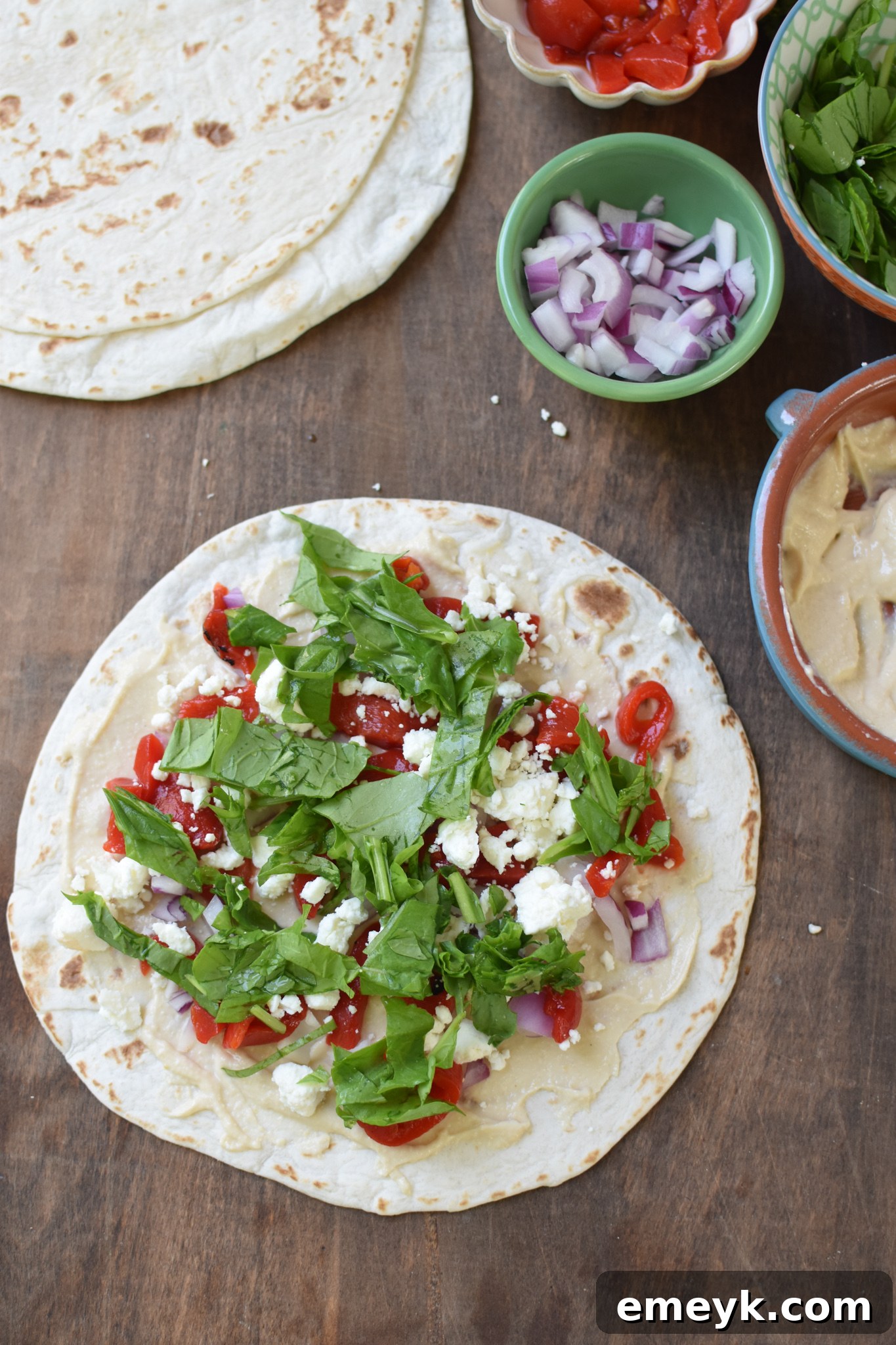A tortilla topped with a vibrant array of Mediterranean ingredients: hummus, roasted red peppers, feta, arugula, red onion, and parsley.