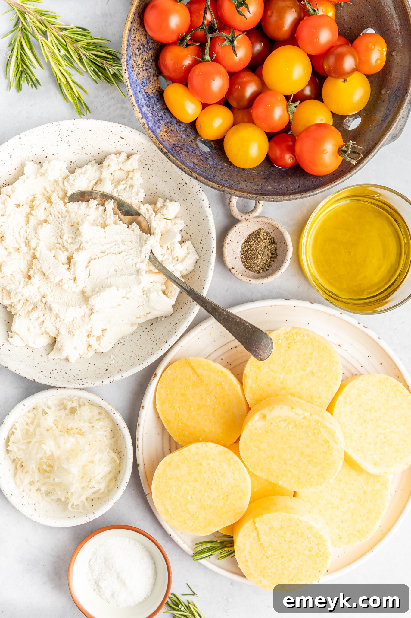 Ingredients for Crispy Polenta Stacks laid out, including polenta tube, cherry tomatoes, fresh rosemary, olive oil, and cheeses.