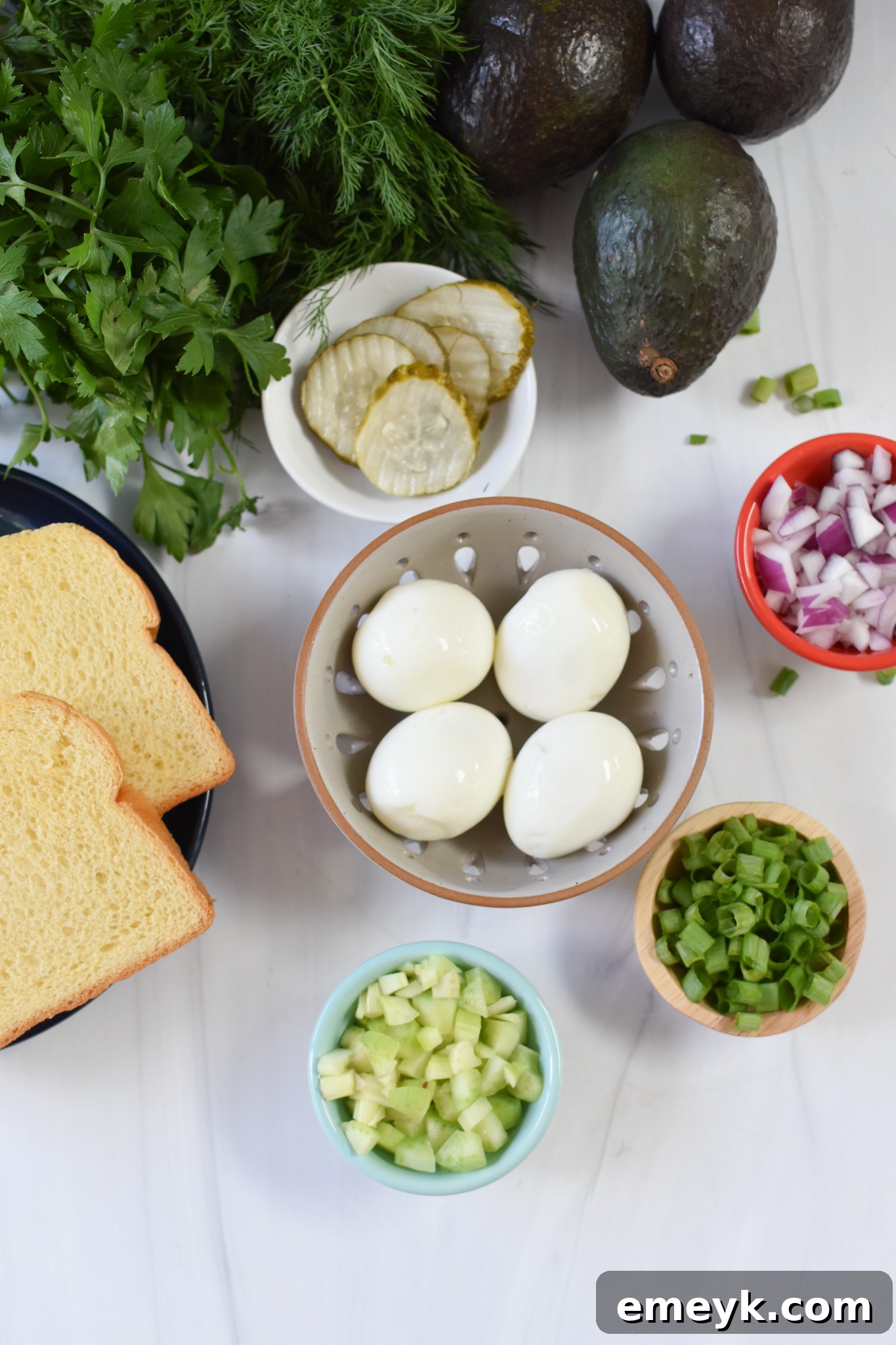 A vibrant display of fresh ingredients for Avocado Egg Salad, including whole eggs, a ripe avocado, crisp pickles, and a medley of herbs and onions, ready for preparation.
