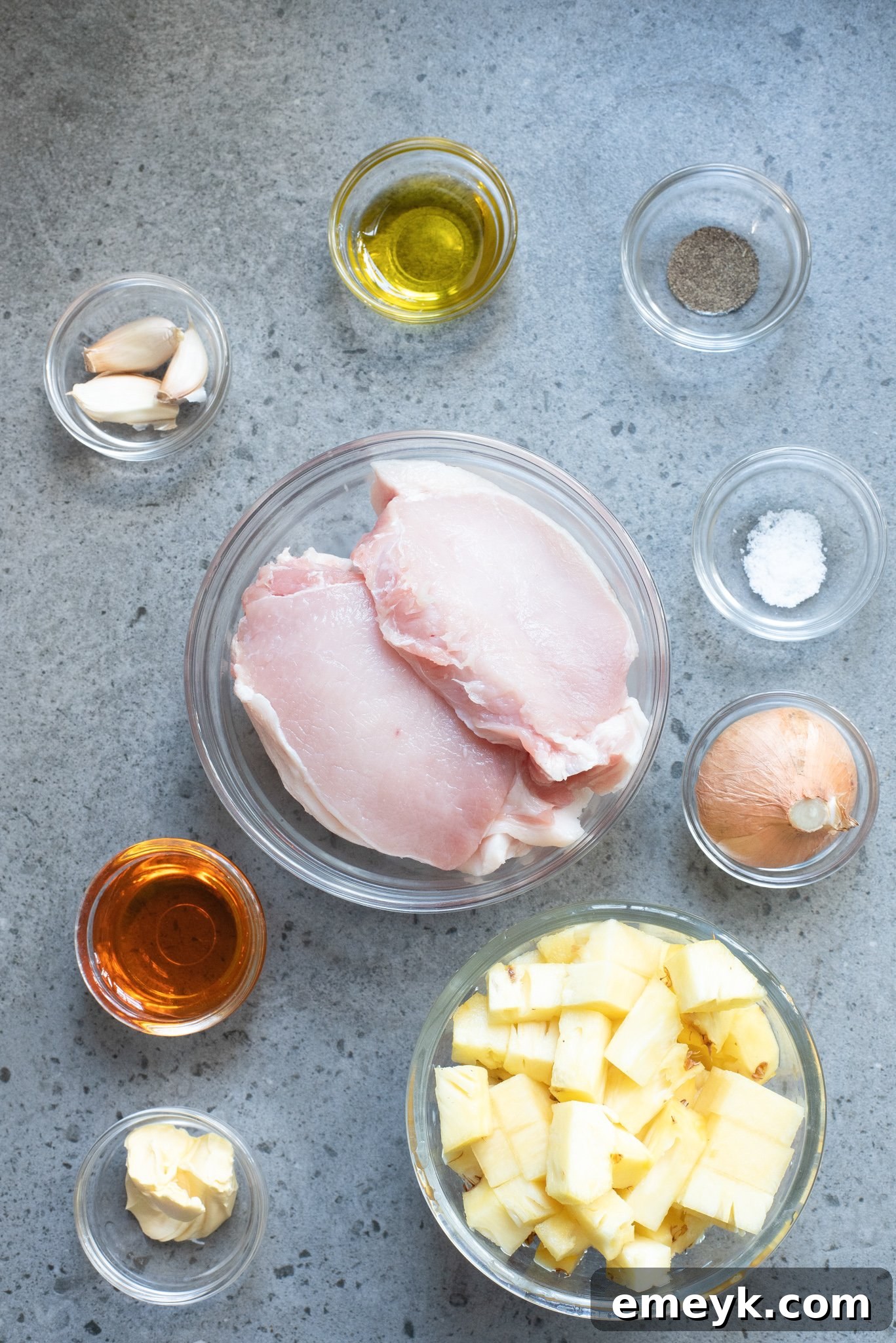 A flat lay photograph showcasing the key ingredients for easy pineapple pork chops: fresh boneless pork chops, a small white onion, garlic cloves, fresh pineapple chunks, a jar of honey, and basic seasonings like salt and pepper.