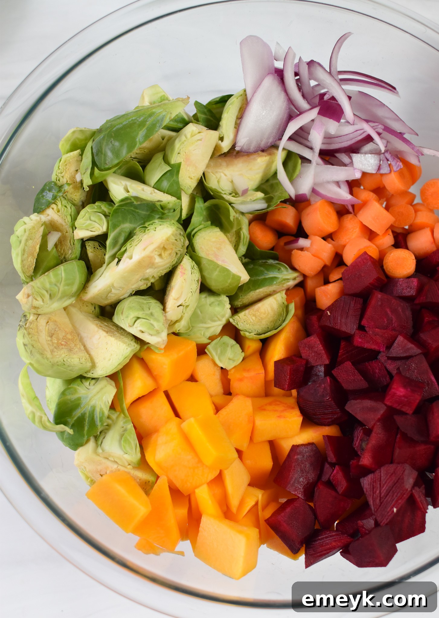 A bowl of fresh, chopped vegetables including butternut squash, Brussels sprouts, beets, red onion, and carrots, ready for roasting.