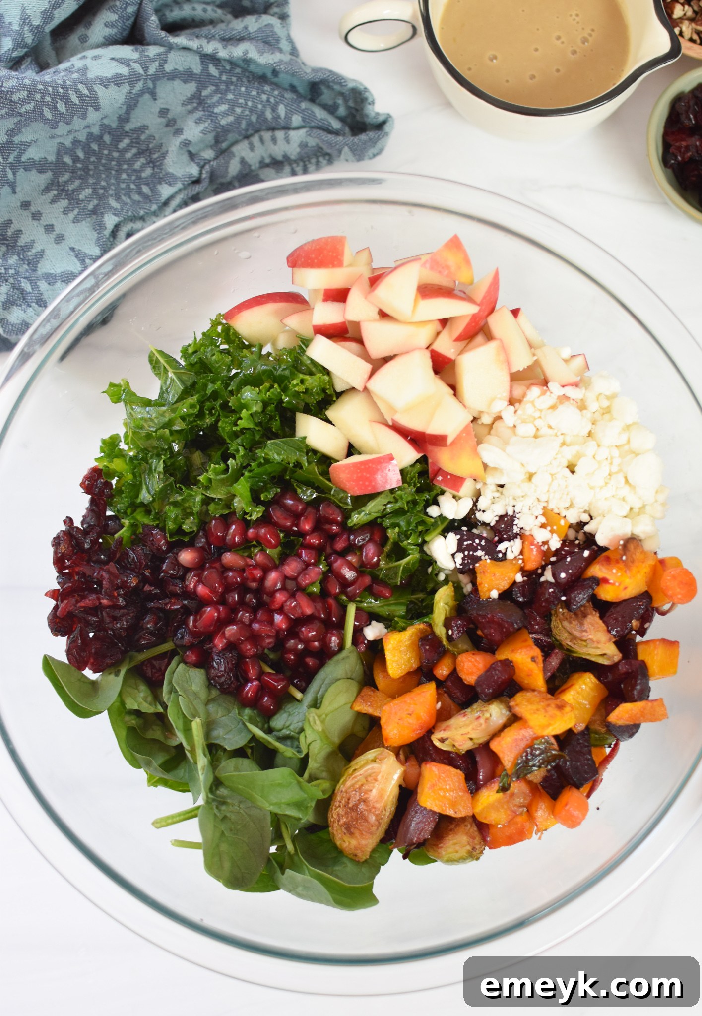 A large mixing bowl filled with fresh salad ingredients: kale, spinach, diced apple, pecans, dried cranberries, pomegranate arils, and goat cheese.