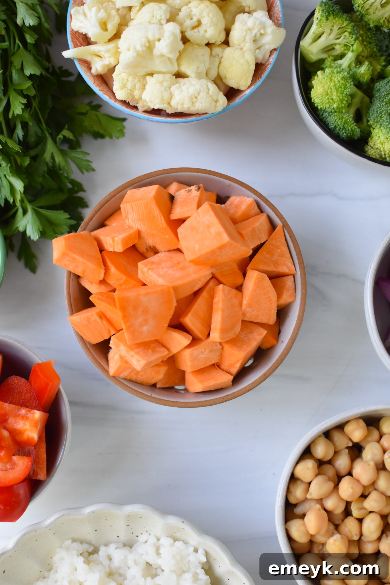 Fresh ingredients for the Sweet Potato Bowl laid out on a table: sweet potatoes, broccoli, cauliflower, carrots, red onion, red bell pepper, chickpeas, rice, greens, and tahini dressing components.