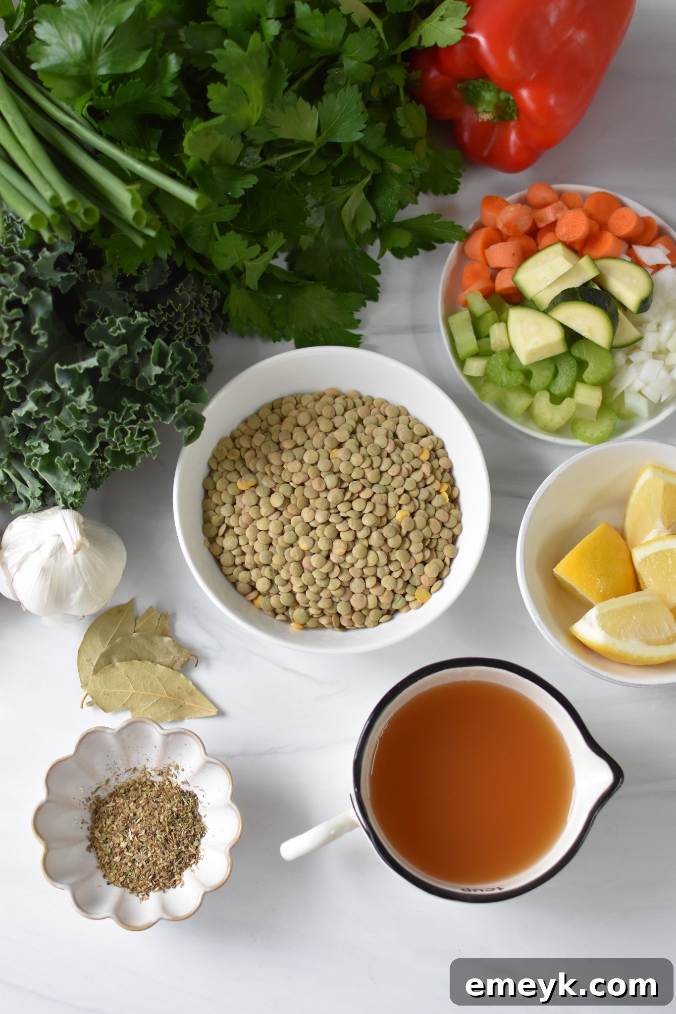A flat lay image showing fresh ingredients for Italian Style Lentil Soup, including green lentils, diced carrots, celery, bell pepper, zucchini, garlic, kale, canned tomatoes, and herbs.