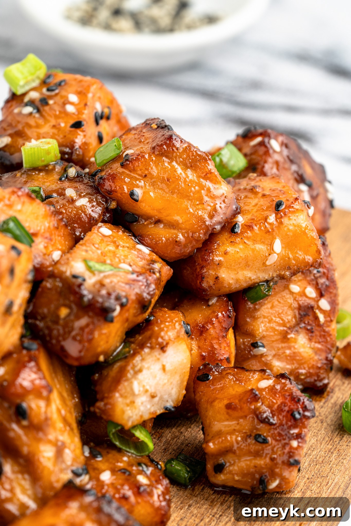 Close-up of glazed salmon cubes garnished with sesame seeds and green onions on a wooden board. A small bowl of sesame seeds is blurred in the background.