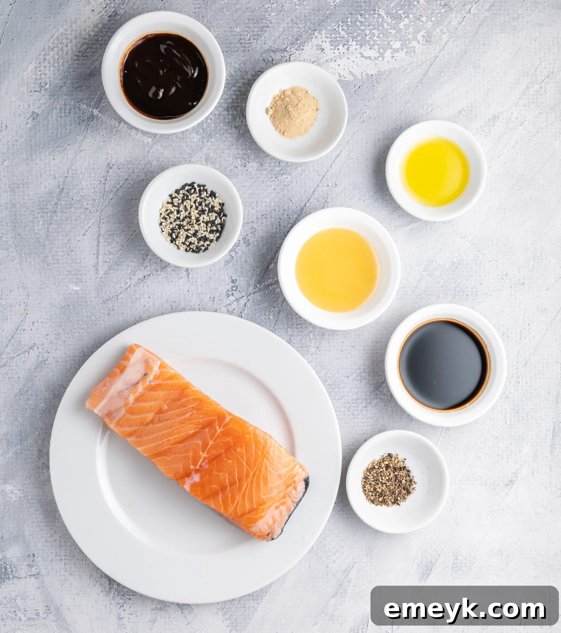 A salmon fillet on a white plate surrounded by small bowls containing various ingredients such as sauce, sesame seeds, oil, garlic powder, and pepper, arranged on a textured gray surface.