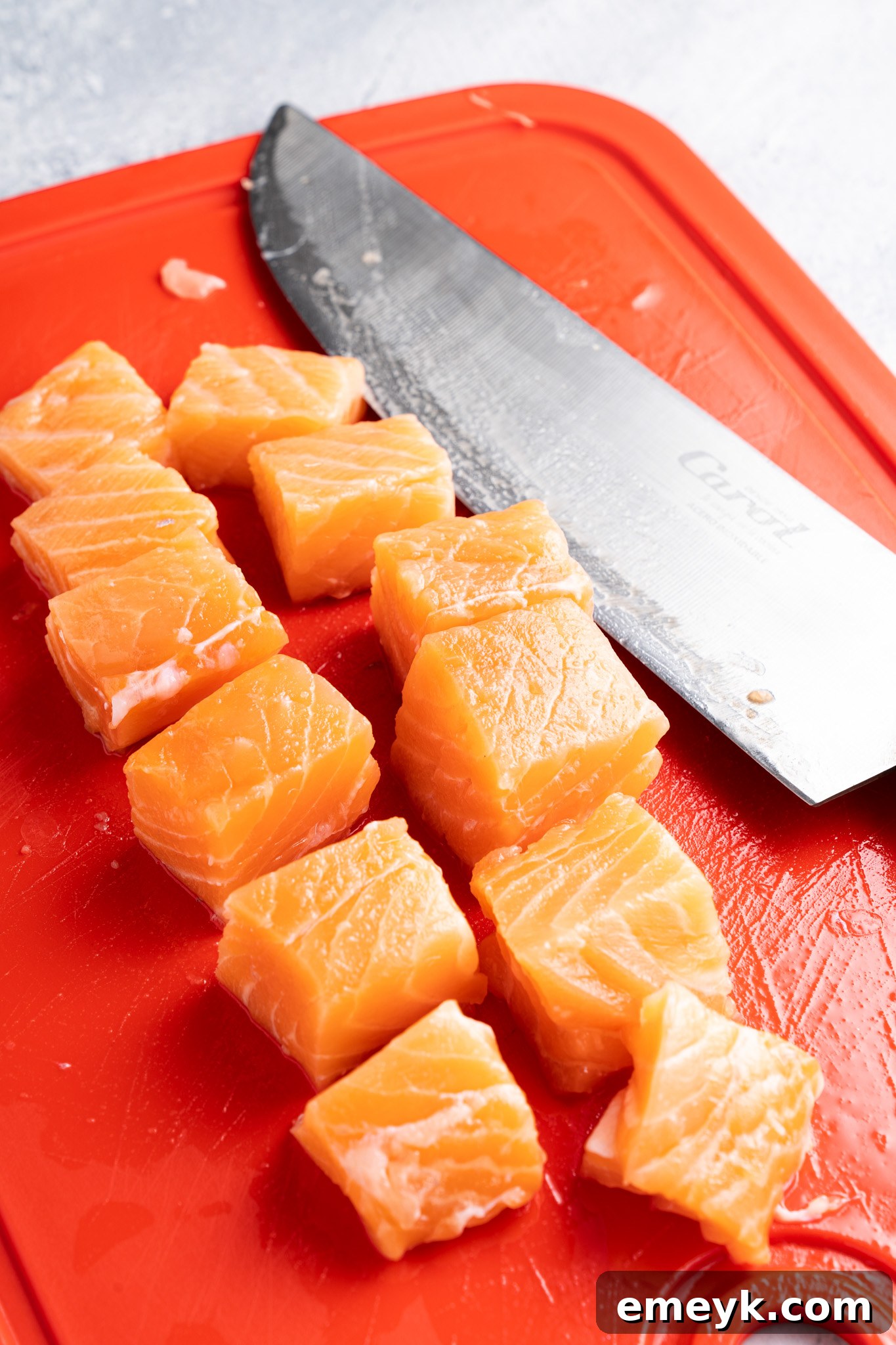 Cubes of fresh salmon are neatly arranged on a bright red cutting board. A large kitchen knife with a shiny blade is positioned next to the salmon pieces. The background has a textured, cool-toned surface.