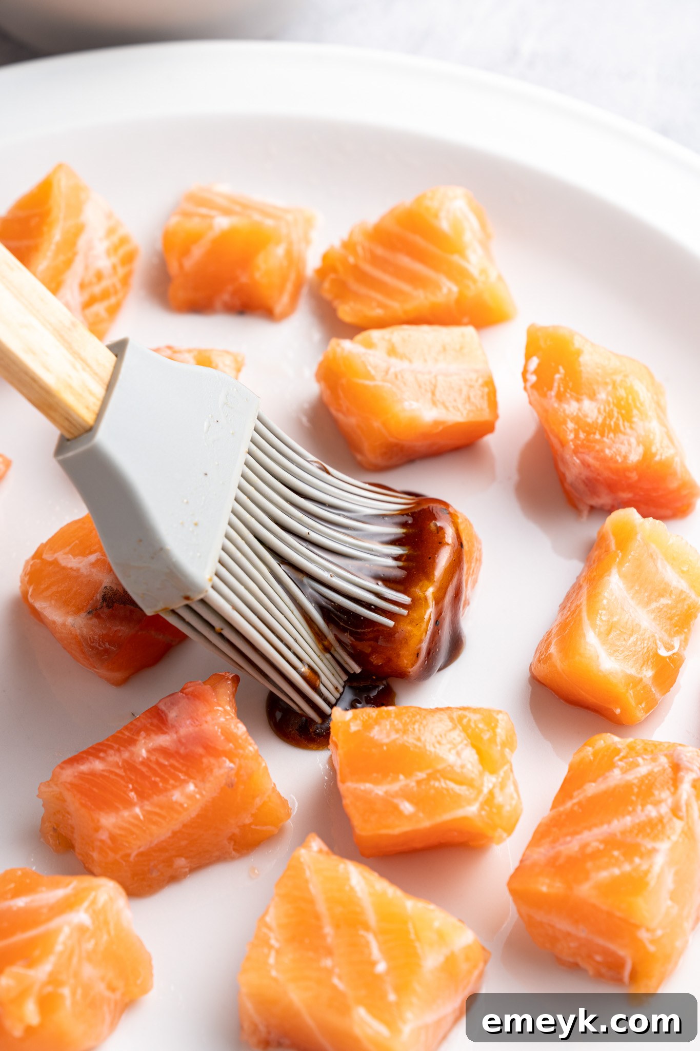 A silicone brush applies a dark marinade to raw salmon cubes on a white plate. The salmon pieces are evenly spaced and glistening, suggesting preparation for cooking. The background is softly blurred.
