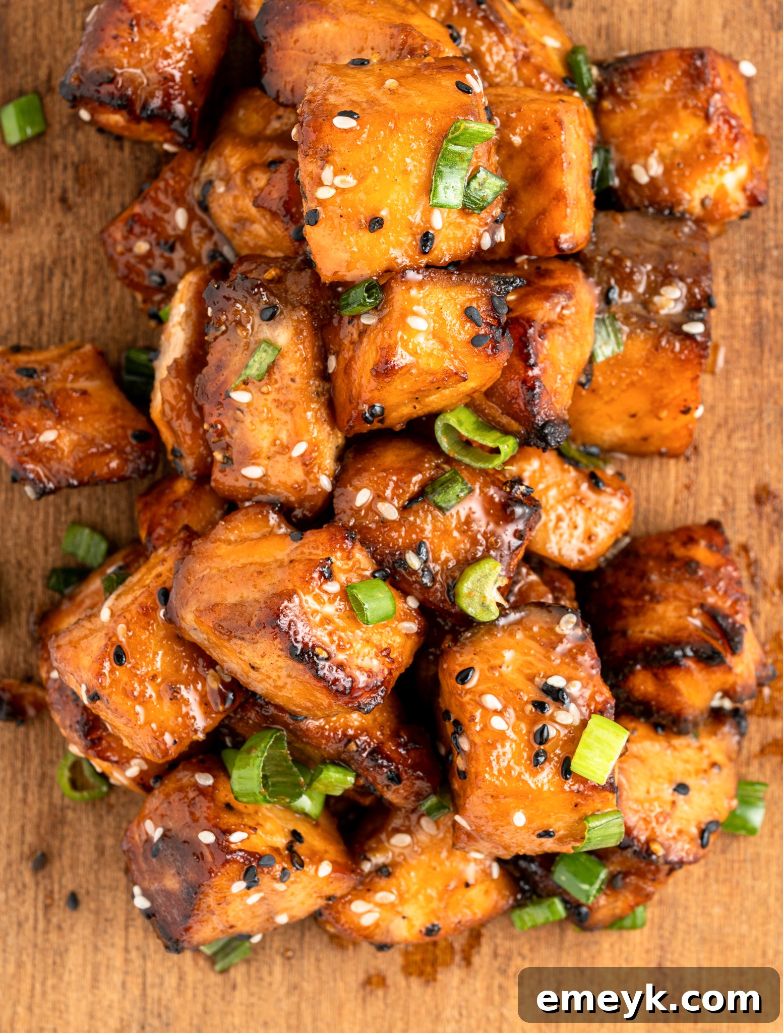 A pile of glazed, air fryer salmon bites topped with sesame seeds and sliced green onions on a wooden surface. The salmon has a caramelized appearance, indicating its well-cooked and flavorful.