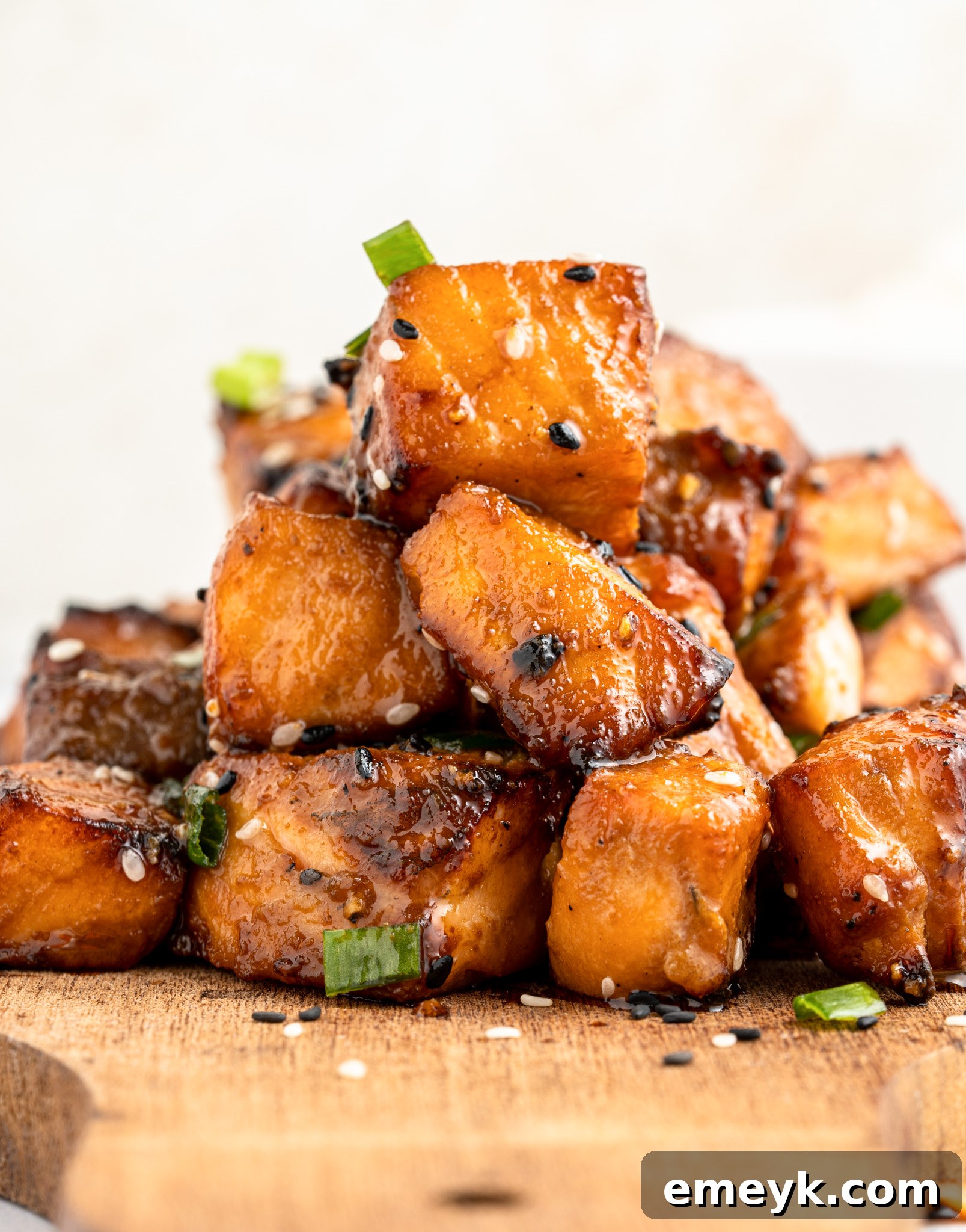 A stack of air fryer salmon bites garnished with sesame seeds and green onions on a wooden board. The pieces are golden-brown, highlighting their caramelized surface.