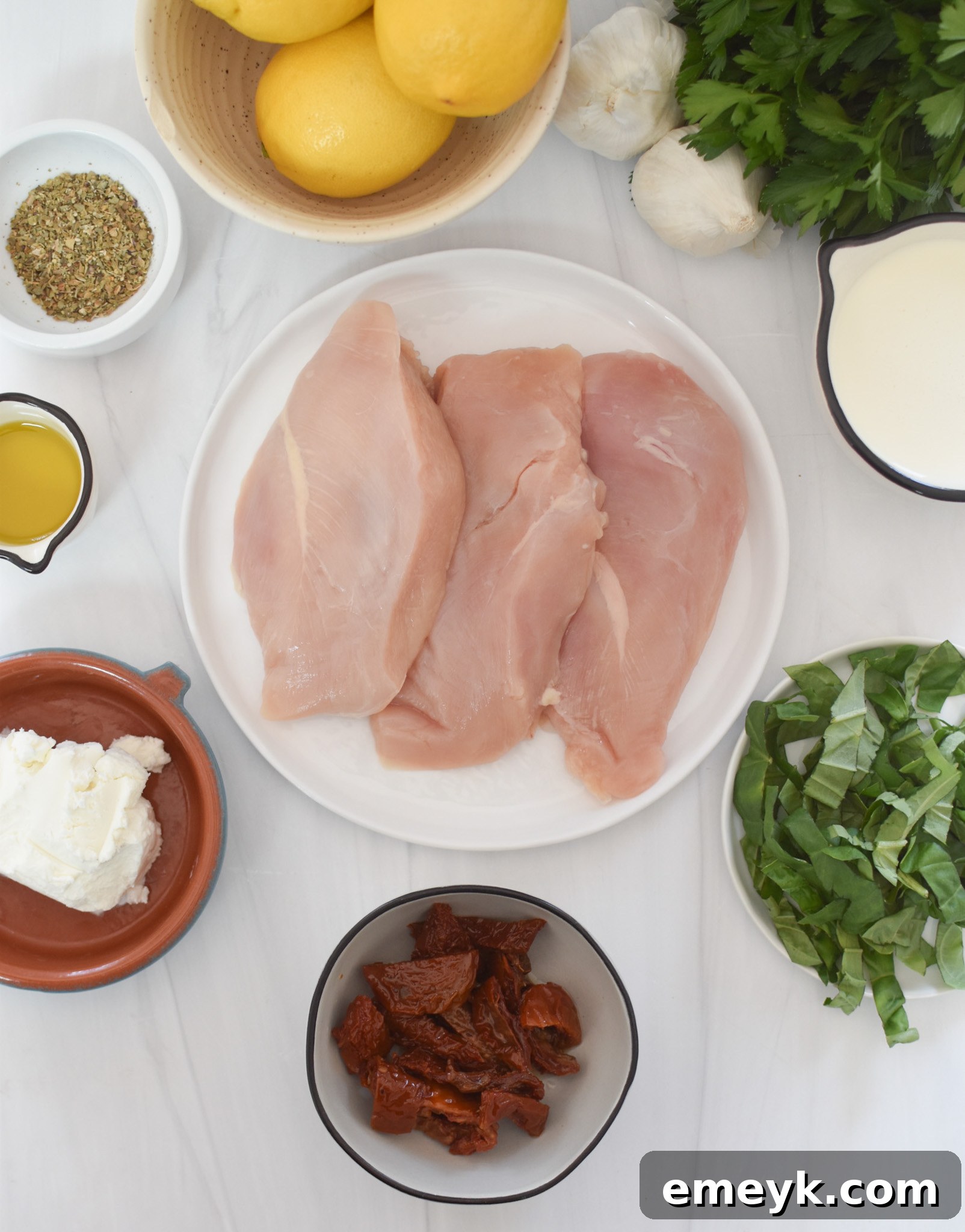 A display of fresh ingredients required for making Chicken Bryan, including chicken breasts, sun-dried tomatoes, goat cheese, basil, and lemons.