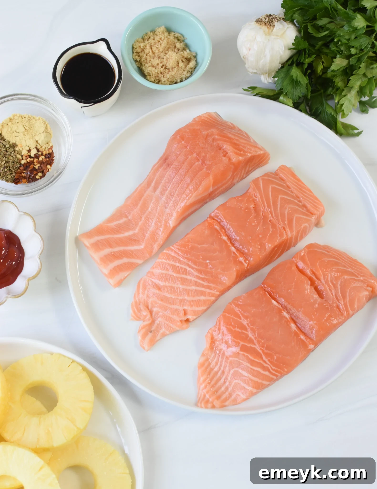 Ingredients for Hawaiian Grilled Salmon laid out on a table, including salmon fillets, fresh pineapple, soy sauce, minced garlic, ginger, brown sugar, ketchup, oregano, red pepper flakes, pineapple juice, and fresh green onions.